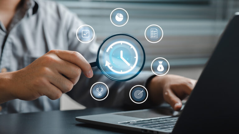 A close-up view of a person using a magnifying glass over a clock icon while working on a laptop, representing business analysis and productivity enhancement.