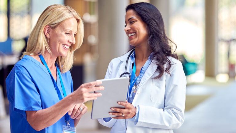 Female Medical Staff In White Coats And Scrubs Use Digital Tablet In Informal Meeting In Hospital