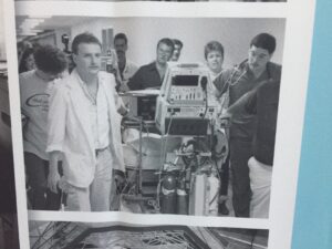 A black and white photo features a young man on the far left with other medical staff wheeling a patient through a hospital hallway.