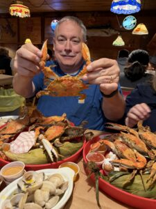 A man with pepper hair smiles while holding up a crab from his seafood boil plate at a restaurant.