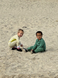 Two young boys play in the sand. One in a yellow striped onsie and the other in a green onsie.